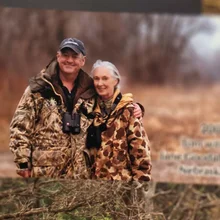 Two people dressed in camouflage jackets standing close together outdoors with binoculars around their necks